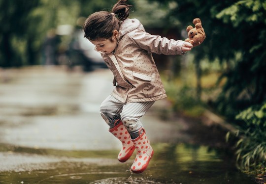 Mädchen mit Regenjacke und Gummistiefeln springt in Pfütze.